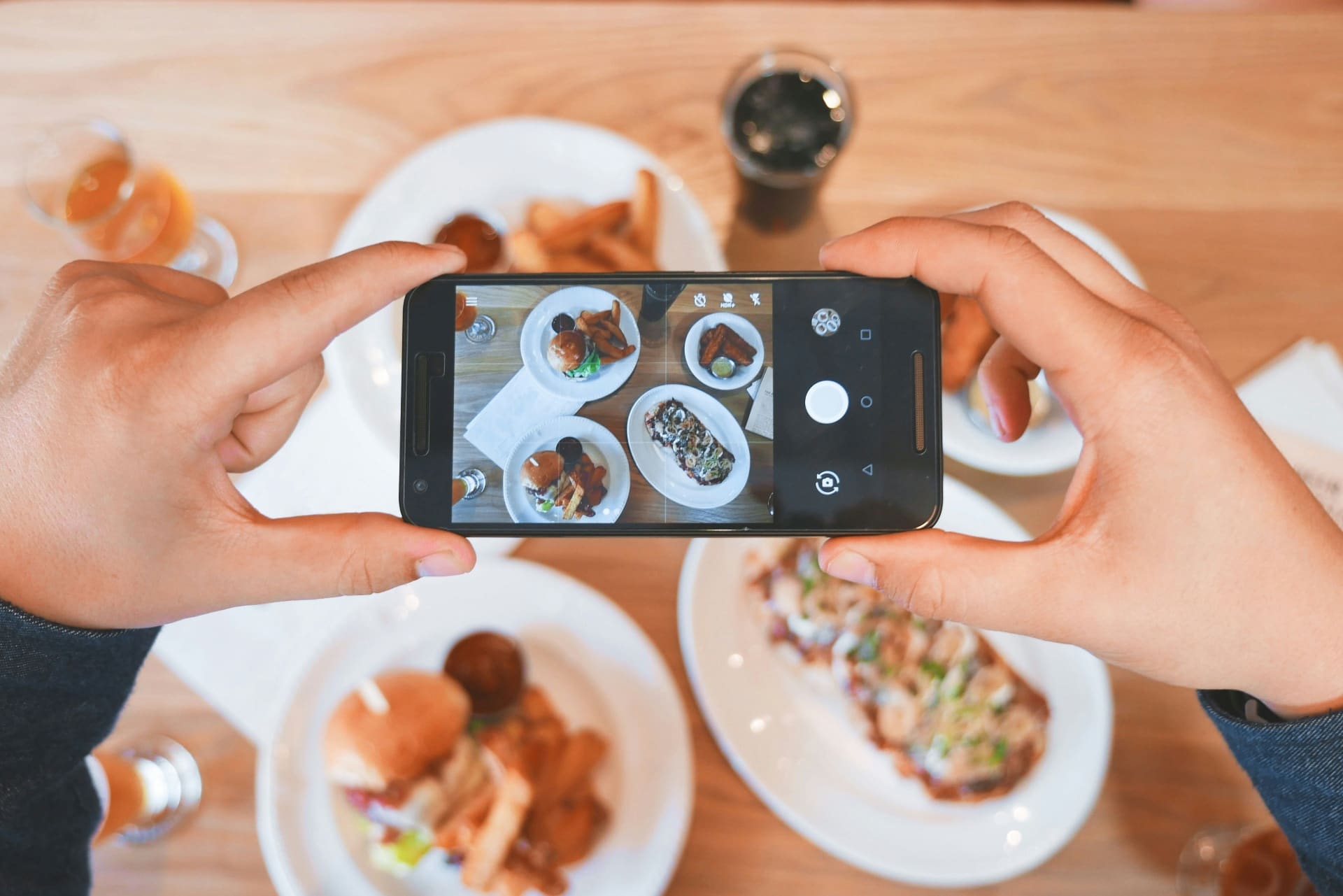 Hand holding a smartphone capturing an overhead shot of burgers, fries and drinks at a restaurant table — behind the scenes hospitality content creation